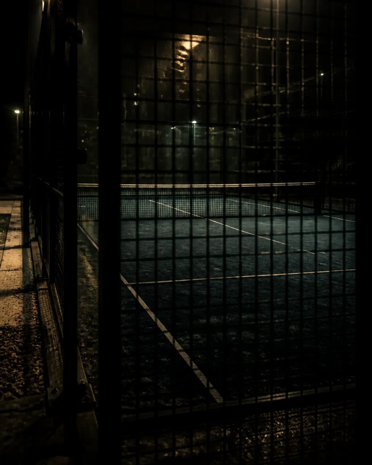 An empty padel court under floodlight at night, seen through the cage